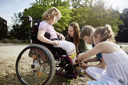 Ein Mädchen sitzt im Rollstuhl, drei weitere Kinder helfen ihr.
