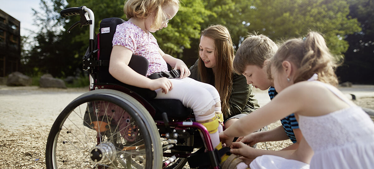 Ein Mädchen sitzt im Rollstuhl und drei weitere Kinder helfen ihr